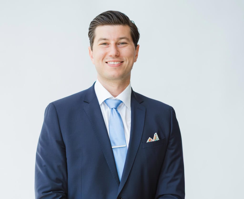 Alex Hughes, Award Winning REALTOR in Victoria, BC , stands smiling at the camera in a blue suit and tie.